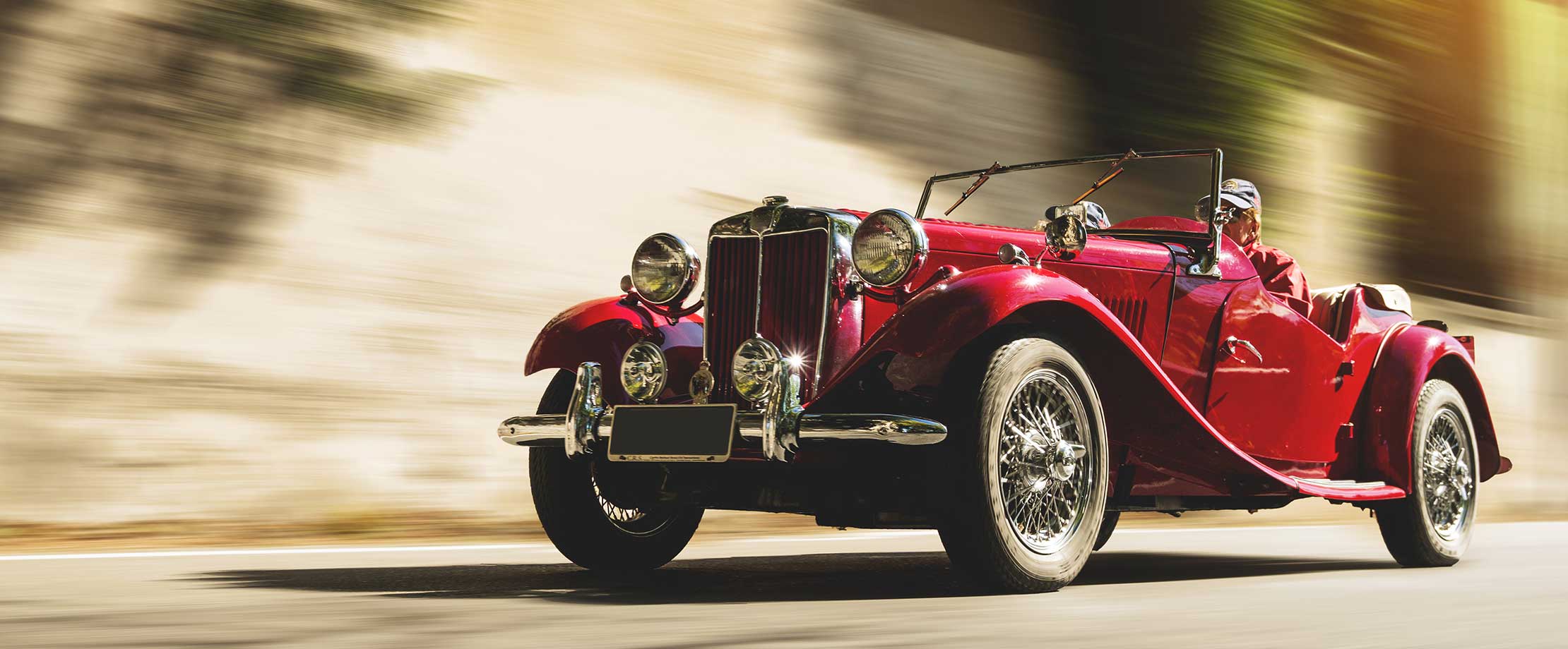 Die beste Versicherung für ihren Oldtimer Ein roter Oldtimer fährt eine Straße entlang. Der Hintergrund ist aufgrund der schnellen Fahrt verschwommen.