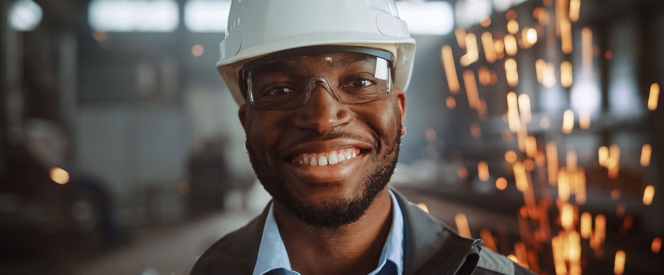 Die beste Absicherung in meinem Beruf Ein Mann steht in einer Werkstatt und lächelt in die Kamera, er trägt einen Schutzhelm und eine Schutzbrille, im Hintergrund fliegen Funken.