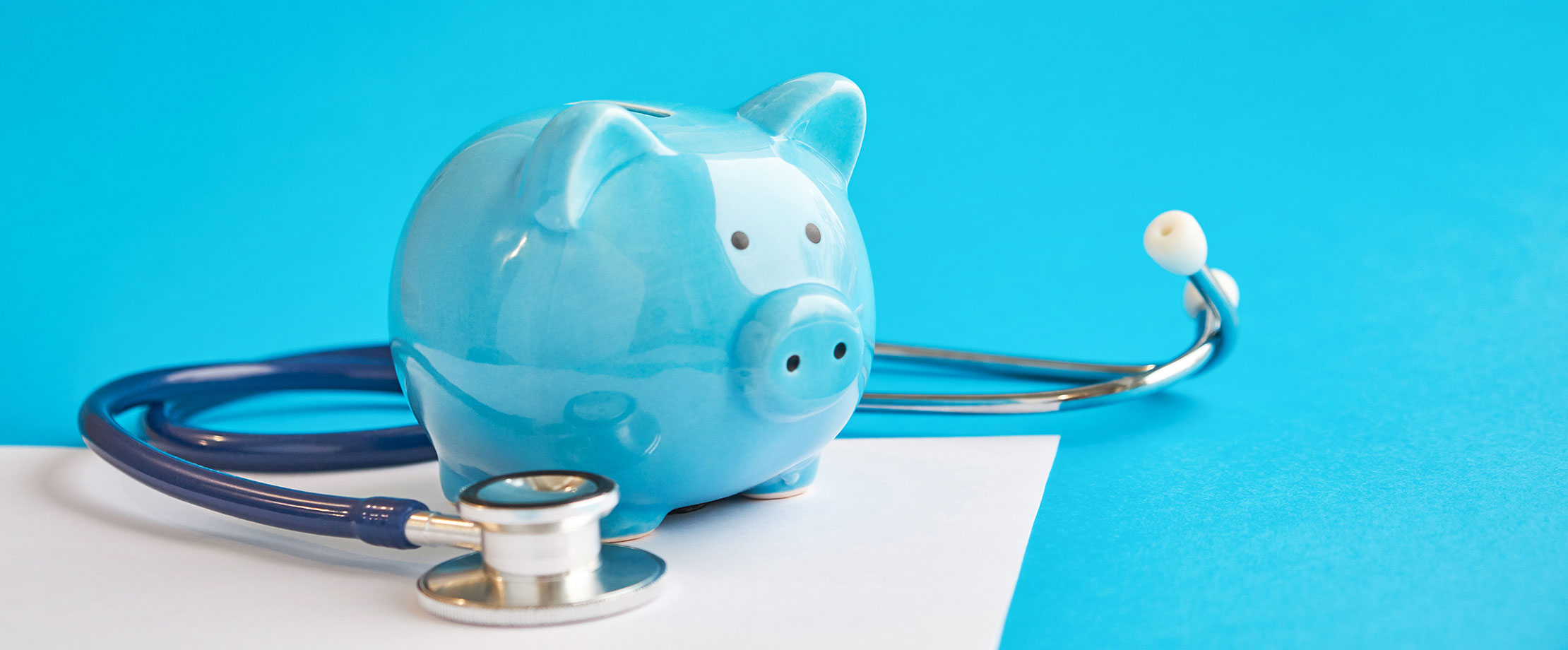 Gesundheitsprüfung Ein blaues Sparschwein steht auf einem weißen Untergrund vor blauem Hintergrund. Naben ihm liegt ein Stethoskop.